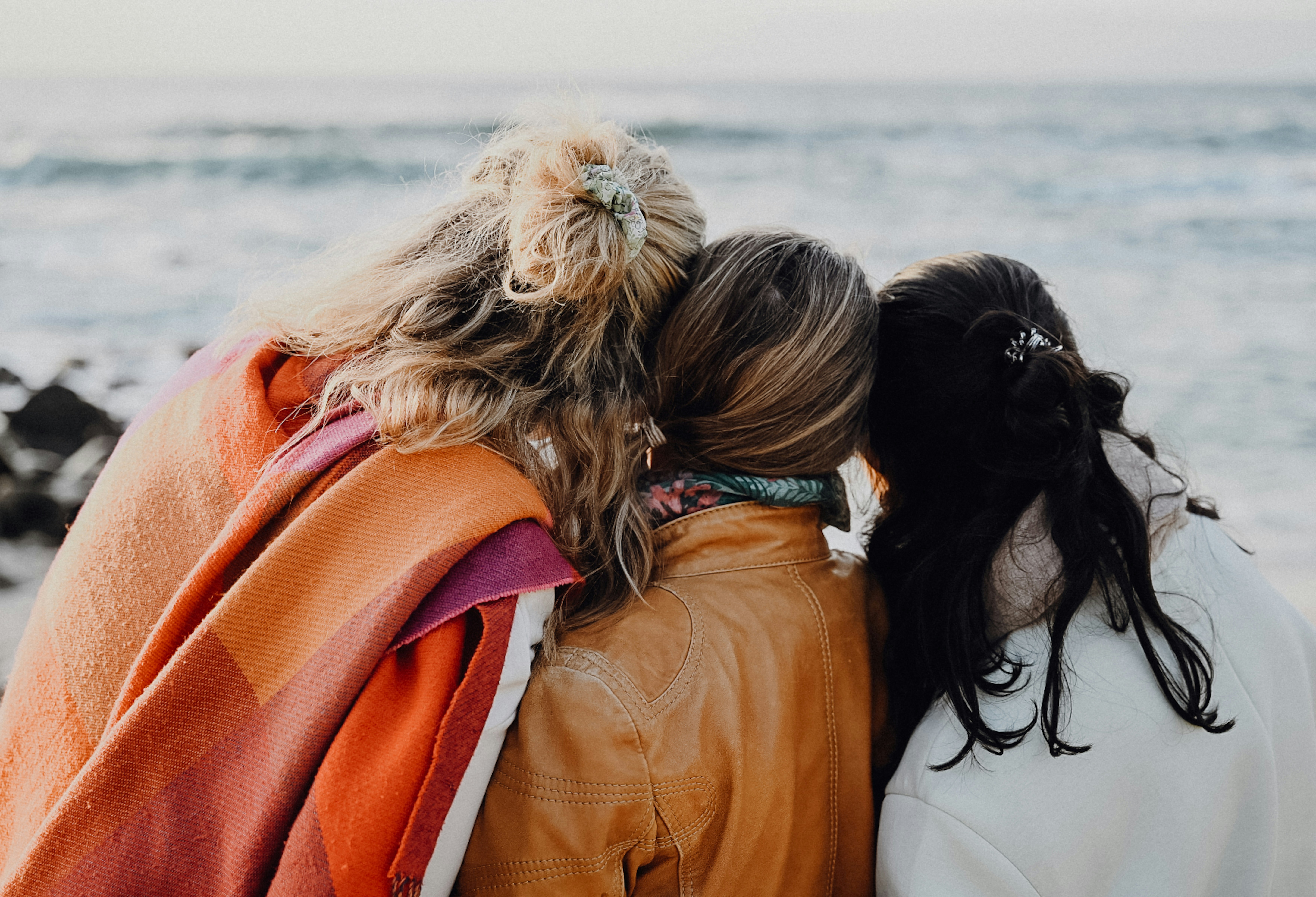 Photo of three women from behind, hugging and in the background is the sea